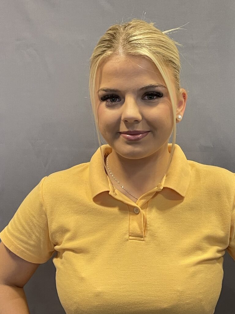 Samira Susami: Junge Frau mit blondem, streng zurückgebundenem Haar, trägt ein gelbes Poloshirt und lächelt dezent vor neutralem grauen Hintergrund