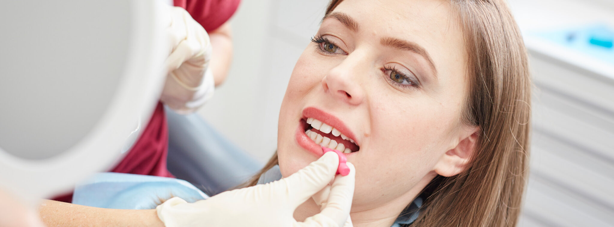 Nahaufnahme einer jungen Frau mit langem dunklen Haar die sich im Handspiegel betrachtet, während ihre Zahnzwischenräume mit einem Interdentalstick behandelt werden.